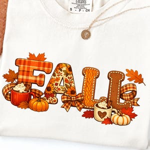 Puede incluir: Una camiseta blanca con un estampado gráfico de la palabra FALL en letras grandes. Las letras están decoradas con cuadros naranjas, girasoles, texturas de punto trenzado y patrones de pan de jengibre, junto a tazas de cacao y hojas otoñales.