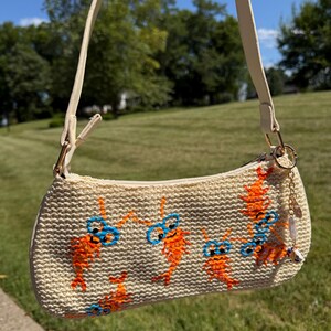 May include: A cream-colored woven shoulder bag featuring a repeating pattern of orange shrimp wearing blue glasses. The bag has a single cream strap, a zipper closure, and a small decorative charm attached to the side ring with gold hardware.