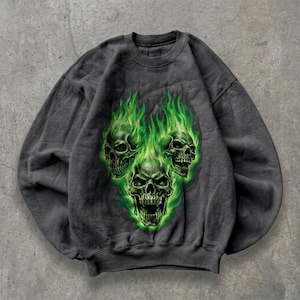 May include: A charcoal gray crewneck sweatshirt featuring a graphic print of three human skulls engulfed in vibrant green flames. The central skull is positioned lower than the two flanking skulls, all depicted with detailed shading and bright green fire.