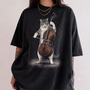 Könnte beinhalten: Ein schwarzes Kurzarm-T-Shirt mit einem Grafikdruck einer grau-weißen getigerten Katze, die auf ihren Hinterbeinen steht und ein braunes hölzernes Cello mit einem Bogen spielt. Die Katze ist mittig auf dem T-Shirt mit Rundhalsausschnitt platziert.