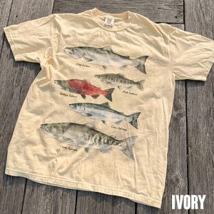 Puede incluir: Una camiseta de manga corta color marfil con un estampado gráfico de cuatro especies de salmón. Las ilustraciones incluyen un salmón real, un salmón rojo, un salmón rosado y un salmón coho, con el salmón keta etiquetado en la parte inferior.