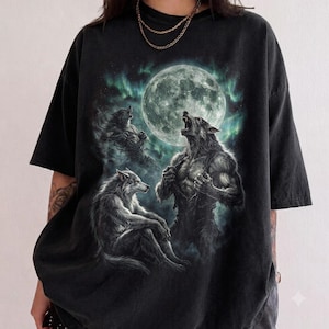 May include: A black short-sleeve t-shirt featuring a graphic print of three wolves and a full moon. The design includes a central werewolf howling, a seated wolf, and a shadowy wolf figure against a backdrop of teal-colored aurora borealis light.