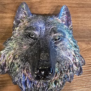 May include: A wolf head wall sculpture featuring a dark, iridescent finish with shimmering highlights of blue, purple, and green. The textured surface captures the detailed fur, pointed ears, and alert eyes of the animal in a realistic, forward-facing pose.