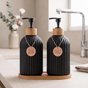 May include: Two matte black ribbed soap dispensers with black pump tops and light wood collars. Each bottle features a round wooden tag hanging from a string, labelled with the words DISH and HAND. The dispensers sit on a matching oval wood tray.