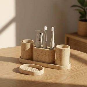 Peut inclure: Un ensemble d'organisation de salle de bain beige moucheté en matériau texturé. L'ensemble comprend un plateau avec deux supports cylindriques, un compartiment central contenant deux brosses à dents électriques blanches et un tube de dentifrice.