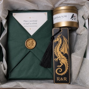 May include: A gift box containing a book wrapped in dark green paper with a gold wax seal, a gold metal candle tin labeled Raven & Rose, a black wooden bookmark featuring a gold dragon illustration and R&R text, and a bundle of dried lavender.