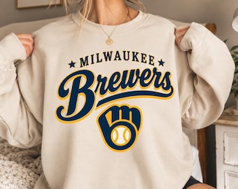 May include: A beige crewneck sweatshirt featuring the text Milwaukee Brewers in dark blue and yellow lettering, accompanied by a dark blue and yellow baseball glove logo with a white baseball in the centre, printed on the front chest.