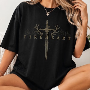 May include: A black short-sleeved t-shirt featuring a gold-coloured graphic print on the chest. The design displays a central sword flanked by deer antlers, surrounded by flame outlines, with the text "FIREHEART" printed in gold capital letters.