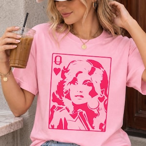 Può includere: Una t-shirt a maniche corte rosa chiaro con una stampa grafica rosa scuro che raffigura il design di una carta da gioco. La grafica mostra il ritratto stilizzato di una donna con capelli voluminosi, un simbolo a cuore e la lettera Q.