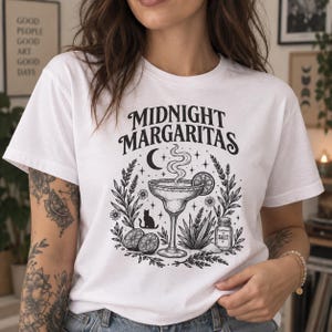 Puede incluir: Una camiseta blanca de manga corta con un estampado gráfico negro con el texto "MIDNIGHT MARGARITAS" sobre una ilustración central de una copa de margarita, una luna creciente, un gato, rodajas de lima, una planta de agave y un salero.
