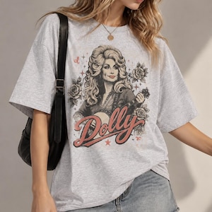 Può includere: Una t-shirt grigio chiaro a maniche corte con una stampa grafica di una donna con voluminosi capelli biondi che suona il banjo. Il design include rose, stelle e la parola Dolly in un carattere rosso corsivo. La maglia ha un girocollo e vestibilità ampia.