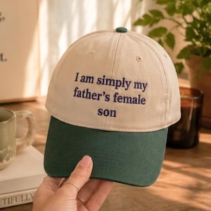 May include: A two-toned baseball cap with a beige crown and a dark green brim. The front of the crown features the embroidered text "I am simply my father's female son" in dark blue lettering. The cap has a button on top and stitched eyelets.