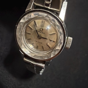 May include: A silver-toned Omega De Ville wristwatch featuring a round, faceted crystal bezel that creates a diamond-like pattern. The watch has a light-colored circular dial with simple hour markers, a metallic link bracelet, and a side winding crown.