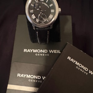 May include: A silver-tone Raymond Weil Geneve wristwatch with a black dial, Roman numeral markers, and a black leather strap. The watch is displayed with its black branded presentation box and an international guarantee booklet.