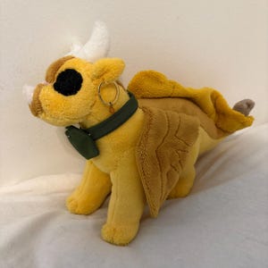 May include: A yellow plush dragon toy with white horns, large black eyes, and a dark green collar featuring a small pouch. The dragon has textured wings, a dorsal fin along its back, and a small gold hoop earring attached to the collar.