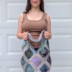 May include: A handmade crochet tote bag with a diamond pattern in shades of gray, blue, and purple. The bag has a long strap and a tassel. The bag is held up by a person.
