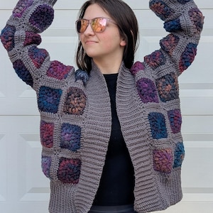May include: A gray, open-front crochet cardigan with colorful square patches on the front and sleeves. The patches feature shades of purple, blue, and red. The cardigan has a ribbed collar and cuffs.