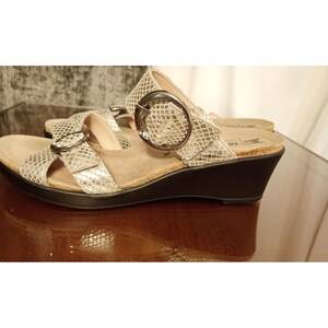 May include: A pair of wedge sandals featuring a metallic silver snakeskin-textured upper with two adjustable straps. Each strap is secured by a large, round, dark metallic buckle. The sandals have a beige contoured footbed and a solid black wedge sole.