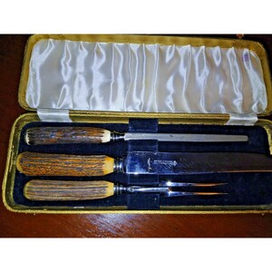Può includere: Un set da intaglio di tre pezzi composto da un coltello, una forchetta e un acciaino, ciascuno con manici in corno marrone testurizzato. Il set è presentato in una custodia dorata con coperchio foderato in raso bianco e base nera.