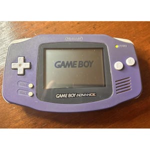 May include: A purple Game Boy Advance handheld gaming console with a white D-pad, A and B buttons, and Start and Select buttons. The screen displays the text GAME BOY, and the console features the Nintendo logo and GAME BOY ADVANCE text.