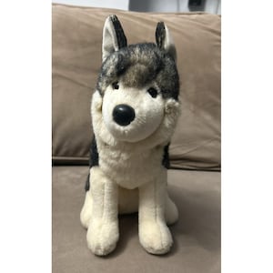 Peut inclure: Une peluche en forme de chien ressemblant à un Husky sibérien avec une fourrure grise, noire et blanche. Le jouet présente des oreilles pointues, un nez noir et des yeux sombres, représenté en position assise avec un pelage doux.