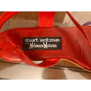 Könnte beinhalten: Eine leuchtend rote Sandale mit einem schwarzen rechteckigen Etikett auf der Innensohle, das den weißen Schriftzug stuart weitzman for Neiman Marcus trägt. Der Schuh hat dünne rote Riemen und einen violetten Akzent an der Seite.