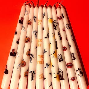 May include: A set of ten white taper candles decorated with various Halloween-themed graphics including orange pumpkins, ghosts, black cats, bats, vampire lips, the Ghostface mask, and the text "TRICK OR TREAT" printed in orange lettering.