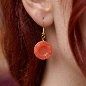 May include: A round, coral-colored earring featuring a recessed center and a textured outer rim. The circular pendant is suspended from a gold-toned metal hook wire, creating a simple drop design that hangs vertically from the earlobe.