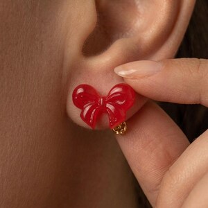 May include: A bright red, glossy bow-shaped stud earring with a smooth, translucent finish. The earring features a small, gold-toned metal backing visible at the base, securely attached to the earlobe, showcasing a simple and clean design.