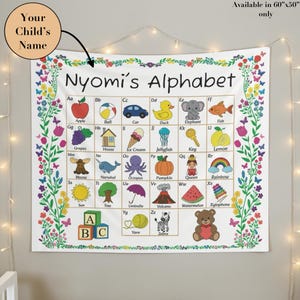 May include: A white fabric wall tapestry featuring the text Nyomi's Alphabet, a grid of letters from A to Z with corresponding colorful illustrations, and a floral border. The tapestry measures 60 inches by 50 inches and includes a personalized name.