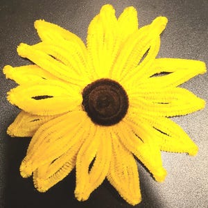 May include: A handcrafted flower made from fuzzy yellow pipe cleaners with a dark brown circular center. The petals are formed by looped yellow chenille stems arranged in a radial pattern to resemble a sunflower, creating a textured appearance.