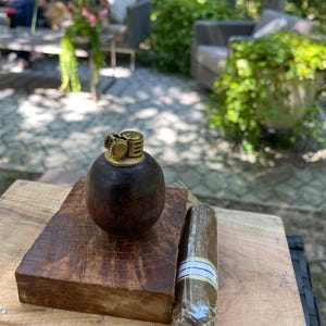 Może przedstawiać: Sferyczna, ciemnobrązowa drewniana zapalniczka ze złotym metalowym mechanizmem zapłonowym stoi na kwadratowym drewnianym klocku obok cygara owiniętego w przezroczystą folię z białą papierową opaską z cienkimi niebieskimi liniami.