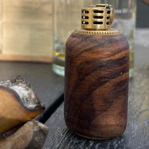 Pode incluir: Um isqueiro cilíndrico com corpo de madeira castanho-escura com veios visíveis e um mecanismo superior em metal dourado. O isqueiro tem um acabamento liso e polido na estrutura de madeira e uma borda serrilhada texturizada.