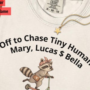 May include: An ivory t-shirt featuring the black text Off to Chase Tiny Humans Mary, Lucas & Bella. Below the text is a graphic of a raccoon wearing a pink bandana and carrying a small bag while riding a scooter. The shirt has a Comfort Colors label.
