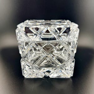 Puede incluir: Una caja cuadrada de cristal transparente con un patrón geométrico facetado en toda su superficie. La tapa y la base están fabricadas en cristal grueso y transparente con surcos profundos que crean un aspecto reflectante y facetado.
