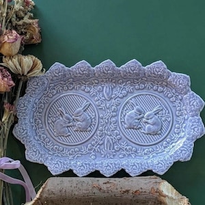 May include: A rectangular lavender ceramic tray with a scalloped edge featuring embossed floral patterns. The center displays two circular medallions, each containing a relief of two rabbits, surrounded by intricate vine and leaf detailing.
