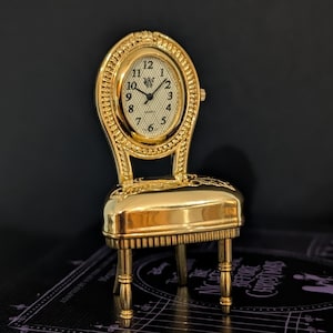 May include: A gold-toned miniature chair-shaped clock with a polished finish. The backrest features an oval clock face with a white textured background, black Arabic numerals, and a small crown on the right side. The seat has embossed details.
