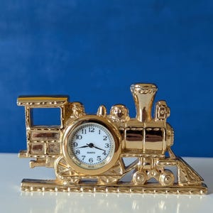 May include: A gold-colored metal train figurine featuring an integrated analog quartz clock. The clock face is white with black numerals and hands. The train design includes a cab, boiler, smokestack, and wheels, all resting on a track base.