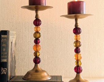 May include: Two brass candle holders with stacked amber and deep red glass spheres on the stems. Each holder features a wide circular base and a flat top plate supporting a lit dark red pillar candle. The holders stand on a dark book.