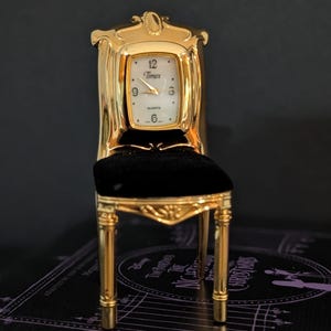 May include: A gold-toned miniature chair clock featuring a white rectangular face with the Timex logo and black numerals. The chair has a black velvet seat, ornate gold-colored frame detailing, and four slender legs, functioning as a quartz timepiece.