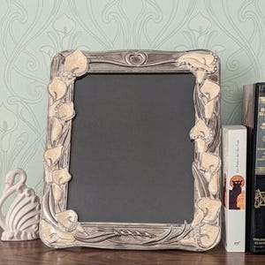 May include: A rectangular silver-toned picture frame featuring a raised relief design of cream-colored calla lilies accented with small clear crystals. The top center of the frame displays a small heart shape, and the center holds a black insert.