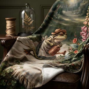 May include: A soft throw blanket featuring a printed illustration of a frog wearing an acorn hat and an apron, holding a copper watering can whilst tending to pink foxglove flowers in a lush garden setting with green clover leaves at the base.