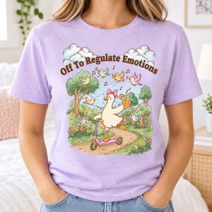 Op de afbeelding: Een lichtpaars t-shirt met een afbeelding van een witte eend met een roze strik op een roze step, terwijl deze een oranje drankje vasthoudt. De tekst "Off To Regulate Emotions" staat erboven, omringd door vogels, bomen en wolken.