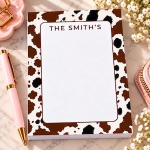 May include: A rectangular notepad featuring a brown and black cow print pattern on the border and a large white central area for writing. The text THE SMITH'S is printed in brown at the top center of the white space within a brown rectangular frame.