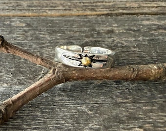 May include: A silver-toned metal ring featuring an open-ended band design. The front of the ring displays a starburst motif with a small, raised gold-toned circular accent at the center, set against a dark, recessed background detail.