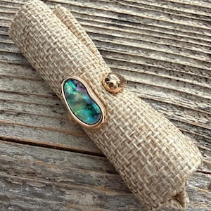 May include: A gold-toned ring featuring an oval-shaped iridescent abalone shell inlay with green, blue, and purple hues. The ring band is smooth and metallic, showcasing a polished finish that complements the natural luster of the shell inlay.