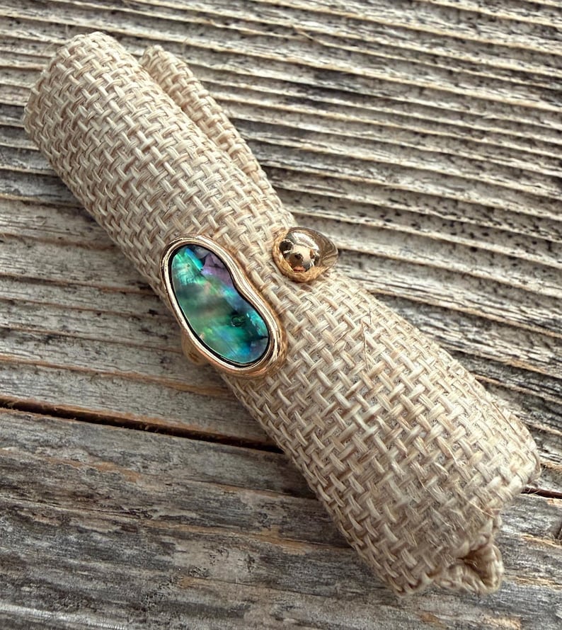 May include: A gold-toned ring featuring an oval-shaped iridescent abalone shell inlay with green, blue, and purple hues. The ring band is smooth and metallic, showcasing a polished finish that complements the natural luster of the shell inlay.