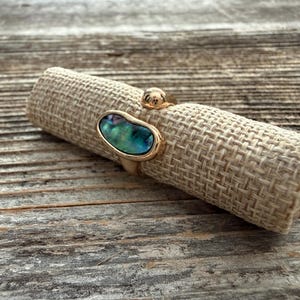 May include: A gold-toned open ring featuring a polished metal band with two distinct ends. One end is capped with a smooth, rounded gold bead, while the other end holds an irregular, oval-shaped inlay of iridescent abalone shell with blue and green hues.