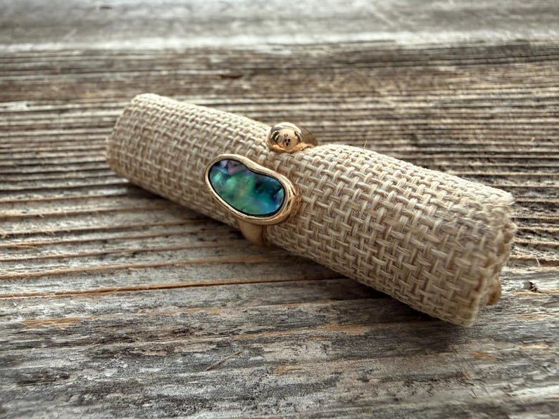 May include: A gold-toned open ring featuring a polished metal band with two distinct ends. One end is capped with a smooth, rounded gold bead, while the other end holds an irregular, oval-shaped inlay of iridescent abalone shell with blue and green hues.