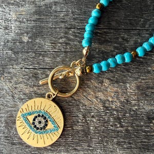 May include: A necklace featuring a strand of round turquoise beads interspersed with small gold-tone metallic beads. The necklace includes a gold-tone toggle clasp and a circular pendant with a stylized eye design, accented by blue and black crystals.